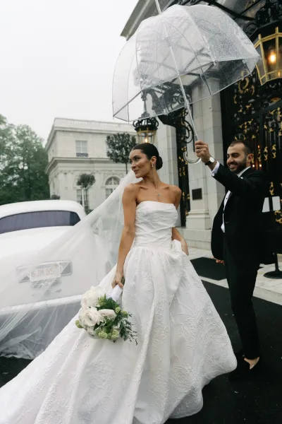 Couple portrait in a rainy wedding photo, sharing a clear umbrella beside an ornate iron gate and stone building facade on wet pavement.