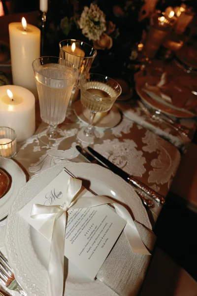 Wedding place setting at a candlelit reception table, a neatly placed menu card atop a damask tablecloth beside a soft floral centerpiece.