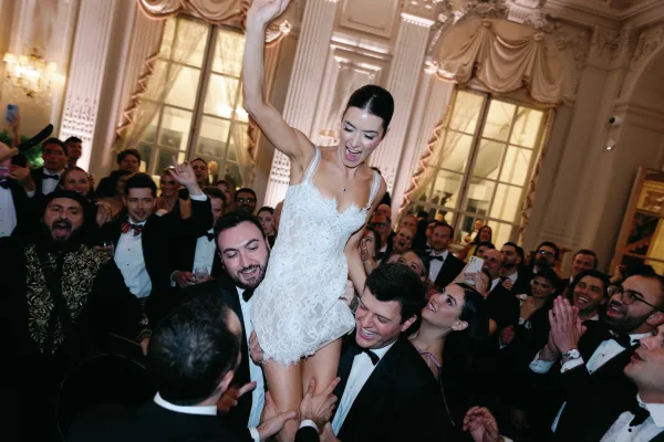 Reception dance moment with the bride lifted in air, wearing a lace mini dress, amid a cheering crowd in a ballroom with tall windows and draped curtains.