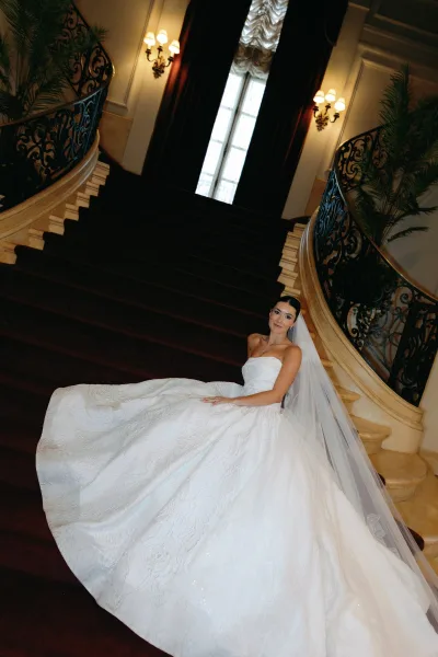 Bridal portrait of a bride in a ball gown wedding dress, long veil trailing as she poses on a red-carpeted grand staircase with wrought iron railing.