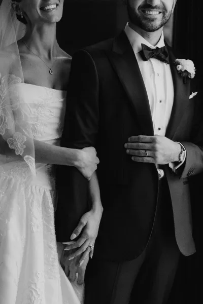 Couple portrait in an arm-in-arm wedding pose, the bride's veil catching light as they smile, framed by a dark interior doorway.