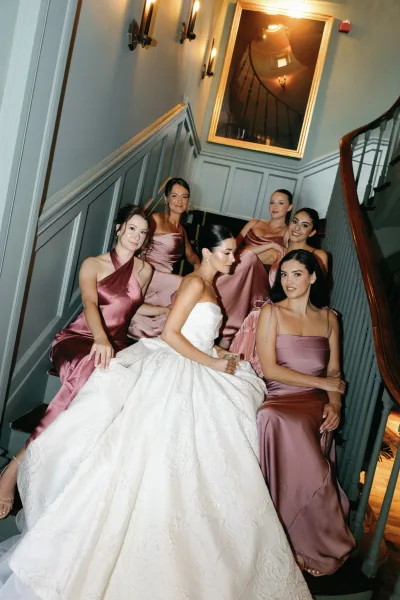 Bridal party portrait of a bride with bridesmaids on an indoor staircase, the bride toasting with a champagne coupe.