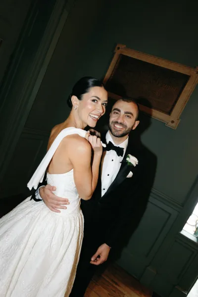 Couple portrait of a bride and groom smiling, his white boutonniere bright as they embrace by window light against painted wall paneling.