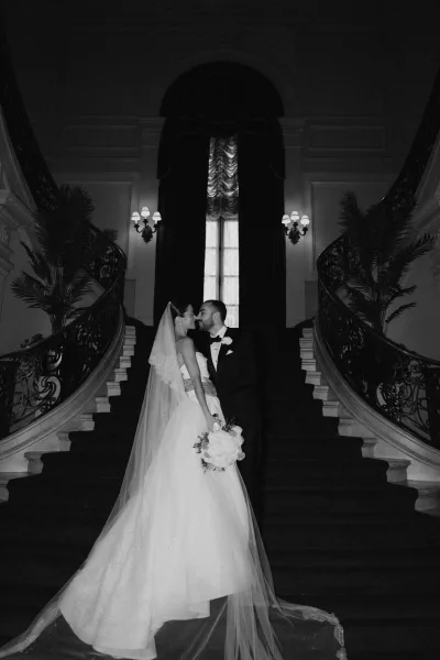 Couple portrait in black and white on a grand staircase, the bride’s veil framed by an ornate railing beneath stately window drapery.
