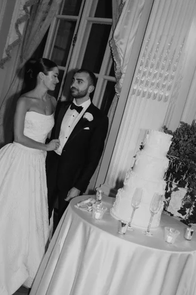 Couple portrait of a bride and groom with cake by a tall window with drapery and greenery, sharing a quiet moment.