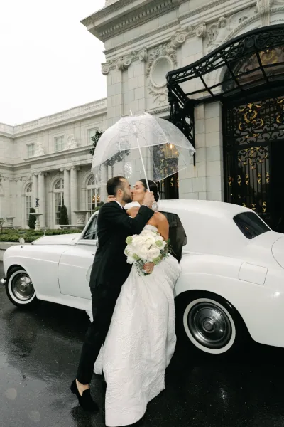 Wedding kiss portrait of a bride and groom under a clear umbrella, with wet pavement and an ornate stone facade behind them.