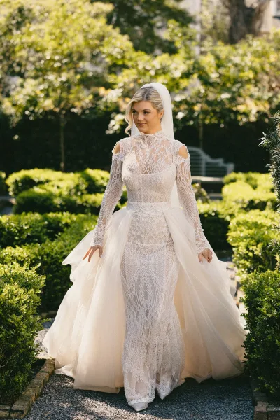 Bridal portrait in a lace wedding dress with cathedral veil, standing on a sunlit gravel path beside manicured garden hedges