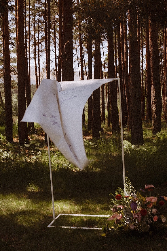 Wedding altar decor with a modern wedding arch, soft fabric draping and ground florals on a sunny grass lawn in a pine forest