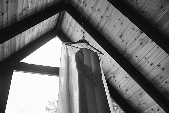 Wedding dress hanging on a hanger, strapless lace bodice softly backlit by window light beneath a wood-beamed ceiling