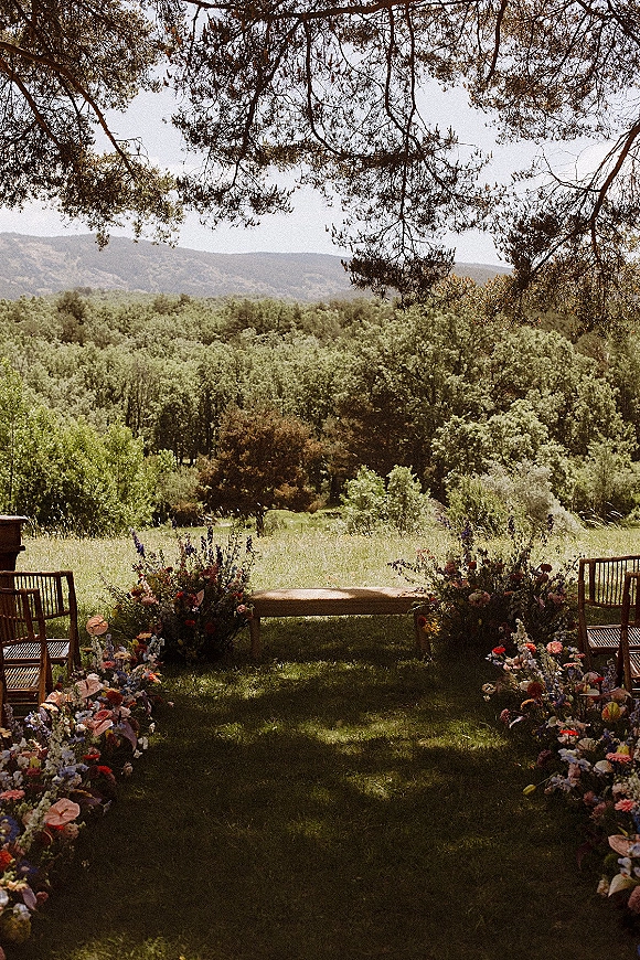Ceremony aisle decor with wildflower ceremony aisle florals lining a grass lawn, wooden chairs and bench under tree branches with hills beyond
