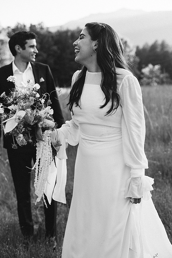 Wedding couple portrait with bride laughing and holding a bouquet beside groom in a suit, walking through a meadow with mountains behind