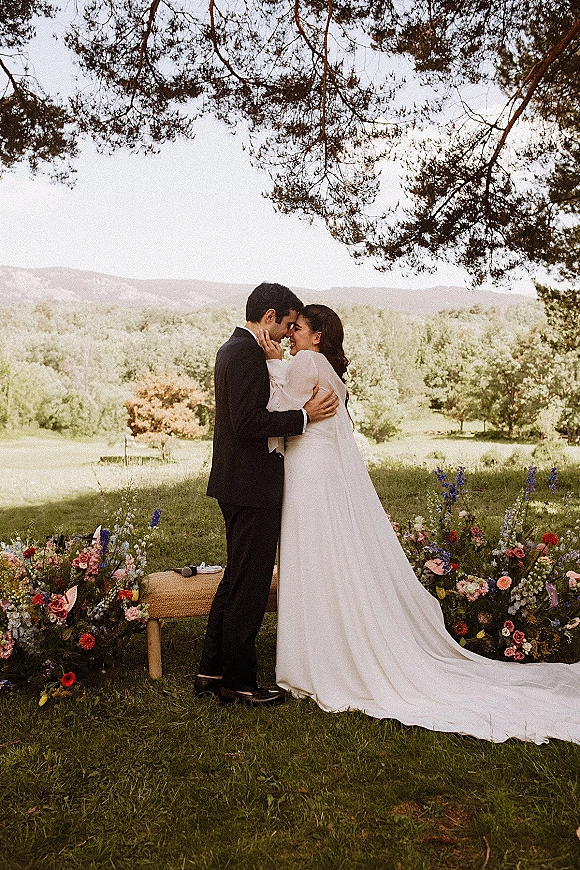 Wedding couple portrait of bride and groom embrace with a forehead touch, her long sleeve gown and train beneath tree branches in a meadow