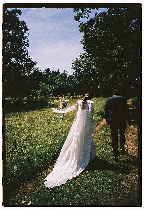Couple walking portrait with wedding dress train, veil, and bouquet, seen from behind on a dirt path through meadow grass and wildflowers