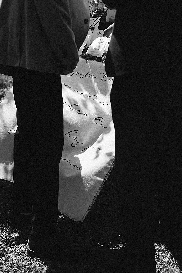 Wedding flat lay featuring a wedding invitation suite with an engagement ring, vow book and calligraphy fabric runner on grass in dappled light