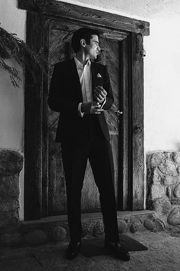 Groom portrait in a black suit with pocket square, standing by a rustic wooden door against a stone and stucco wall backdrop