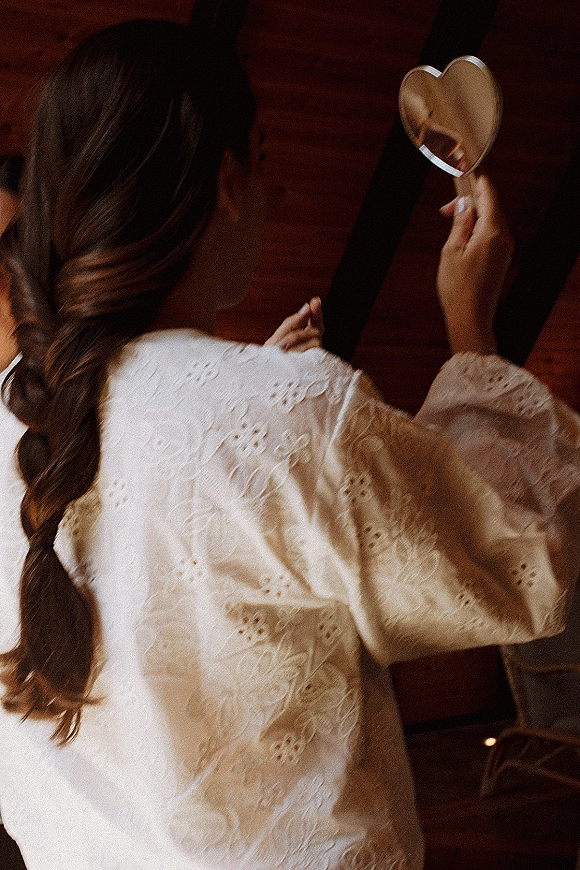 Bridal getting ready as bride in lace robe holds a heart hand mirror, showing braided hair from behind in window light by wood wall