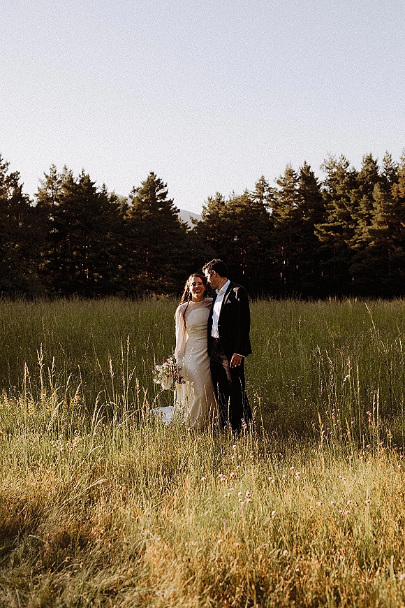 Couple portrait of bride in long sleeve dress and veil holding a white bouquet beside groom in black tux in a wildflower field at golden hour