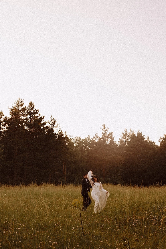 Couple portrait of bride in a wedding dress and veil with groom in tuxedo walking through a wildflower meadow at sunset light
