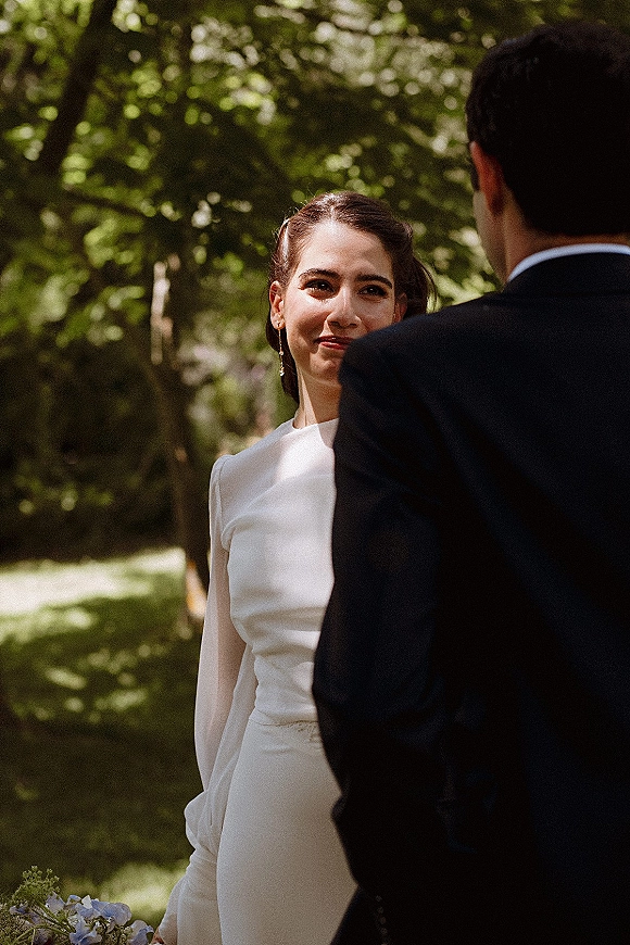 First look moment as bride in long-sleeve wedding dress smiles at groom in black suit, holding bouquet in sunlit forest clearing
