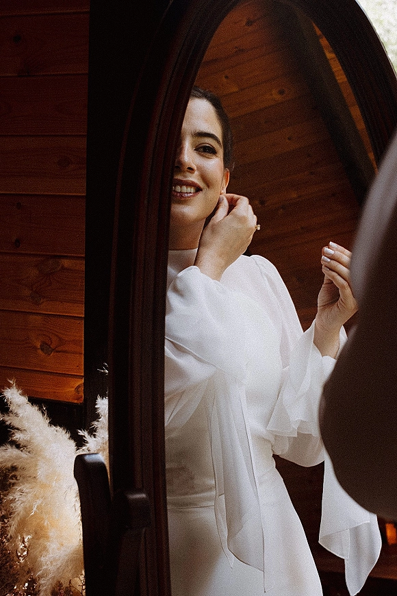 Bridal portrait of a bride getting ready in a high-neck long-sleeve dress, reflected in a mirror with pampas grass by wood paneling