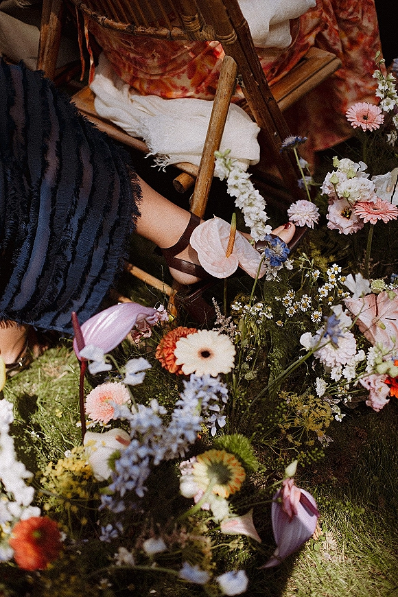 Wedding shoe detail with ankle strap heels beside a ruffled dress hem on a bamboo chair over a grass lawn with garden flowers