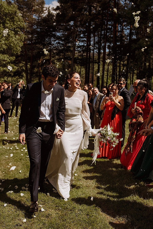 Wedding recessional as newlyweds walk hand in hand while guests toss flower petals, bride laughing with bouquet on a pine-lined lawn