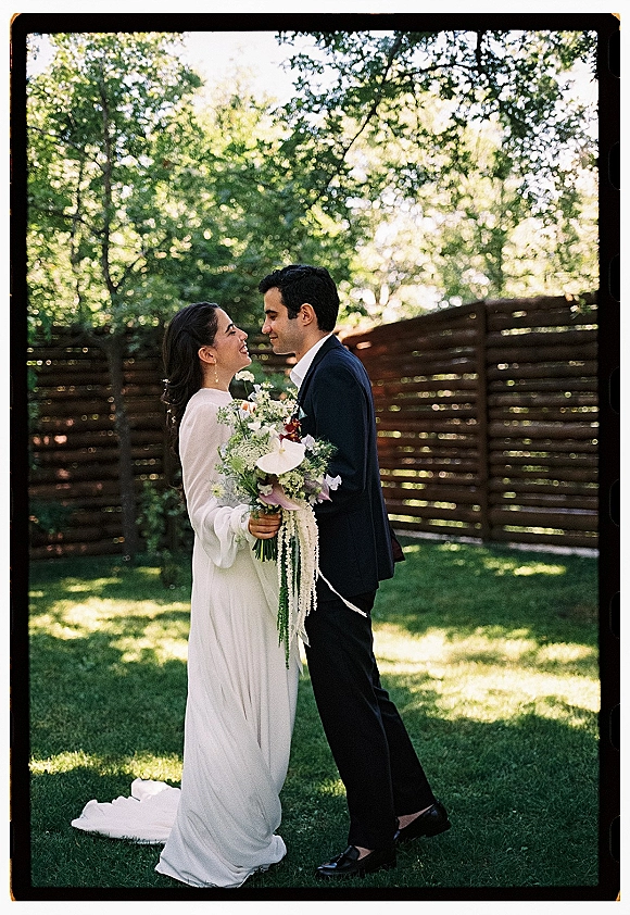 Couple portrait of bride and groom facing each other, bride holding bouquet in sunlight on a garden lawn by a wooden fence