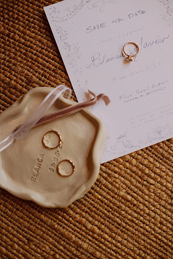 Wedding rings and gold wedding bands beside a solitaire engagement ring in a dish, styled with satin ribbon on a rattan surface