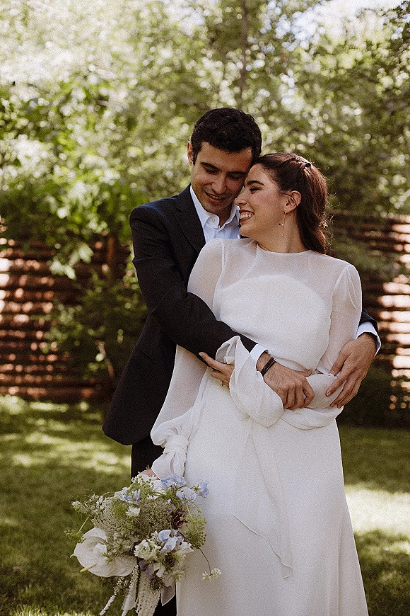 Couple portrait of bride and groom embrace, her white gown and bouquet with ribbon, in dappled sunlight by garden trees and fence