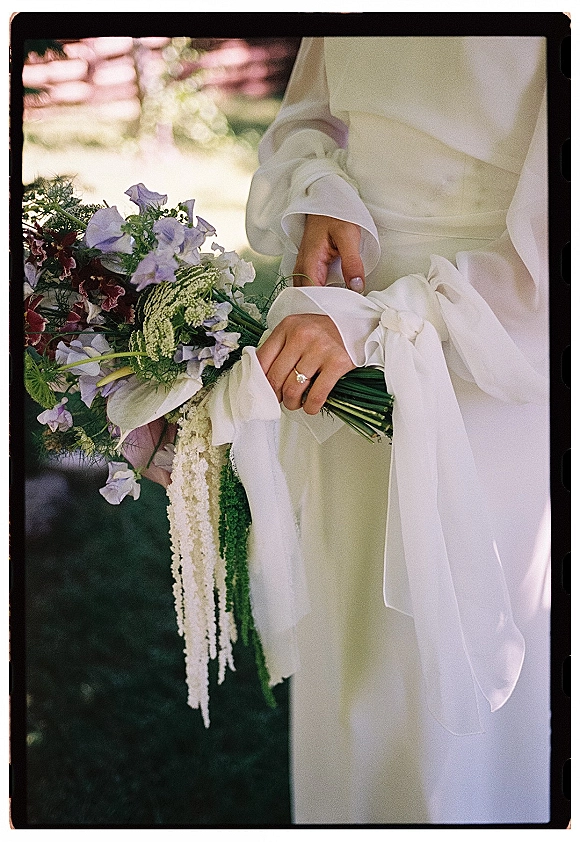 Bridal bouquet with wildflower bridal bouquet blooms tied with a white satin bow, held in ringed hands, trailing ribbons in sunny greenery bokeh
