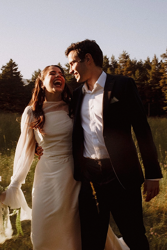 Couple portrait of bride and groom laughing as they walk through a golden meadow with wildflowers and pine-covered hills at golden hour