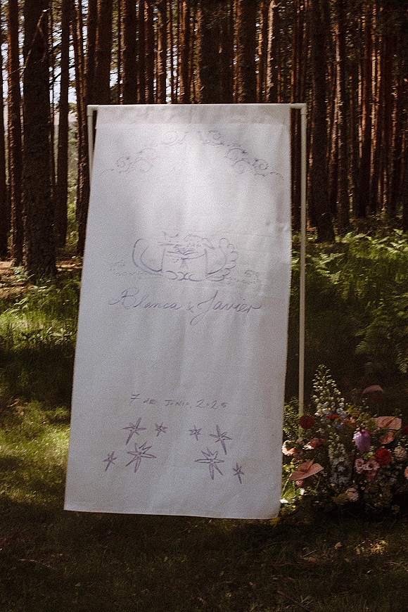 Wedding welcome sign on fabric in a wooden frame with floral arrangement and greenery, set in a sunlit pine forest with ferns