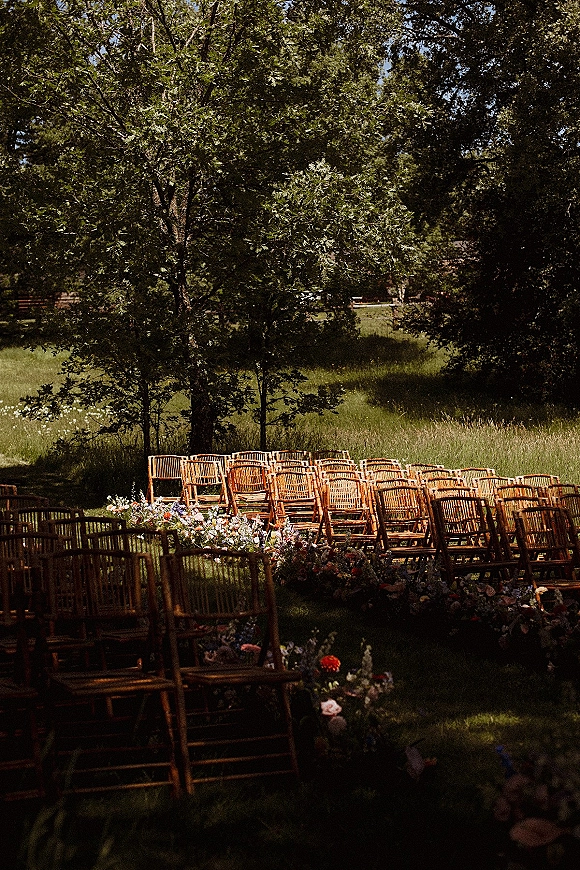 Ceremony seating with wood chairs lined along an aisle of low floral ground arrangements on a grassy meadow beneath trees