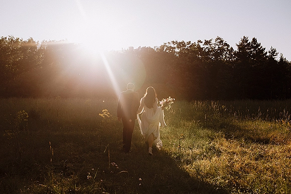 Couple portrait of bride and groom walking away, holding hands in tall meadow grass at sunset, bride holding bouquet and dress train