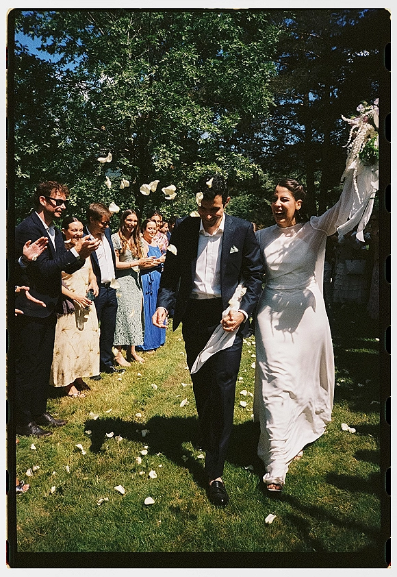 Wedding recessional as bride and groom walk hand in hand, bride raising bouquet with ribbon streamers while guests toss rose petals in garden