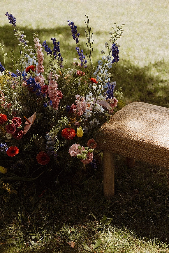 Wedding ceremony florals and aisle flower arrangement in wild roses, delphinium, and tulips beside a woven rattan bench on sunlit grass lawn