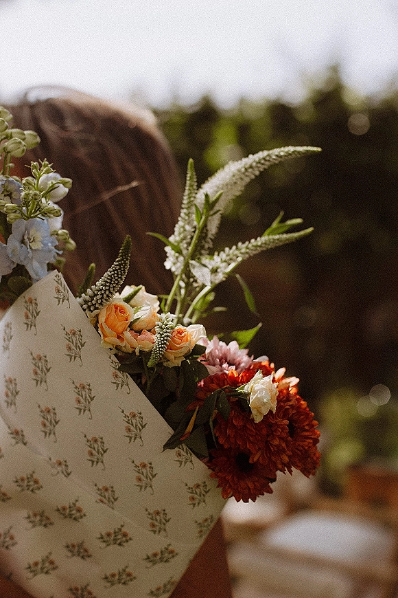 Wedding bouquet with orange rose bouquet blooms, red dahlia, white flowers and greenery wrapped in floral paper against sunlit bokeh greenery