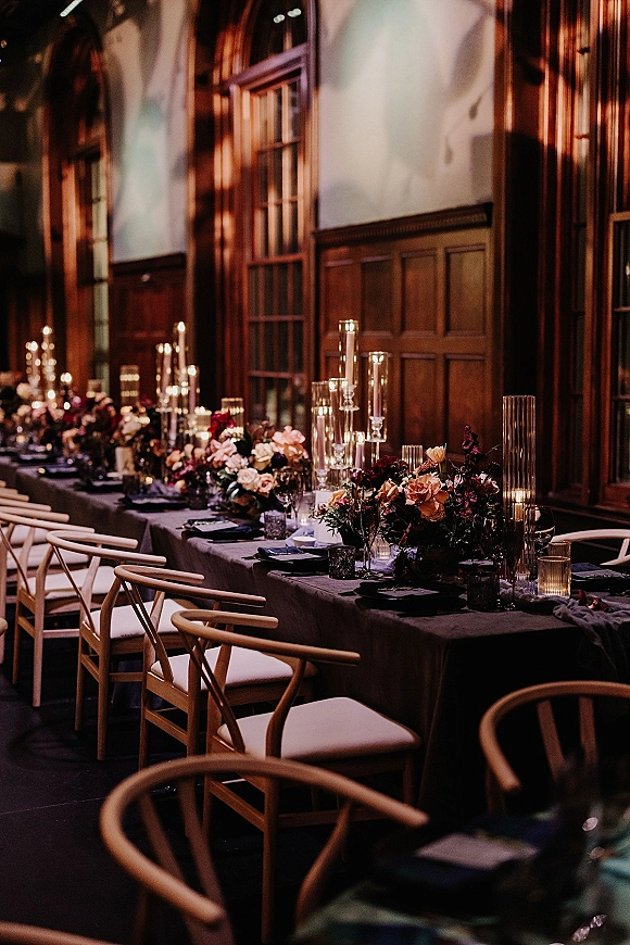 Reception tablescape on a long banquet table wedding with gray linens, floral centerpieces, tall glass candles, and place cards in a wood-paneled hall
