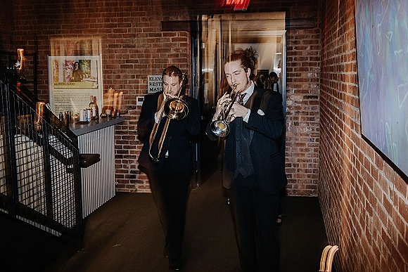 Wedding band of live wedding musicians walking through a brick hallway by a bar, playing trombone and trumpet in black suits