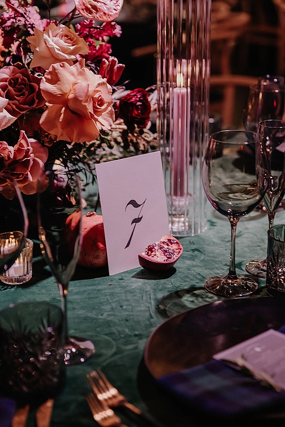 Reception tablescape with an emerald green tablecloth, rose centerpiece, taper candle, pomegranate, and place cards in dim lighting