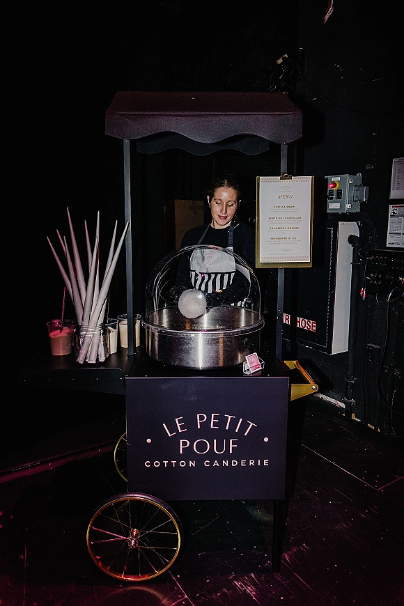 Cotton candy cart wedding cotton candy station with machine, menu sign, and paper cones under a canopy in a dark industrial venue