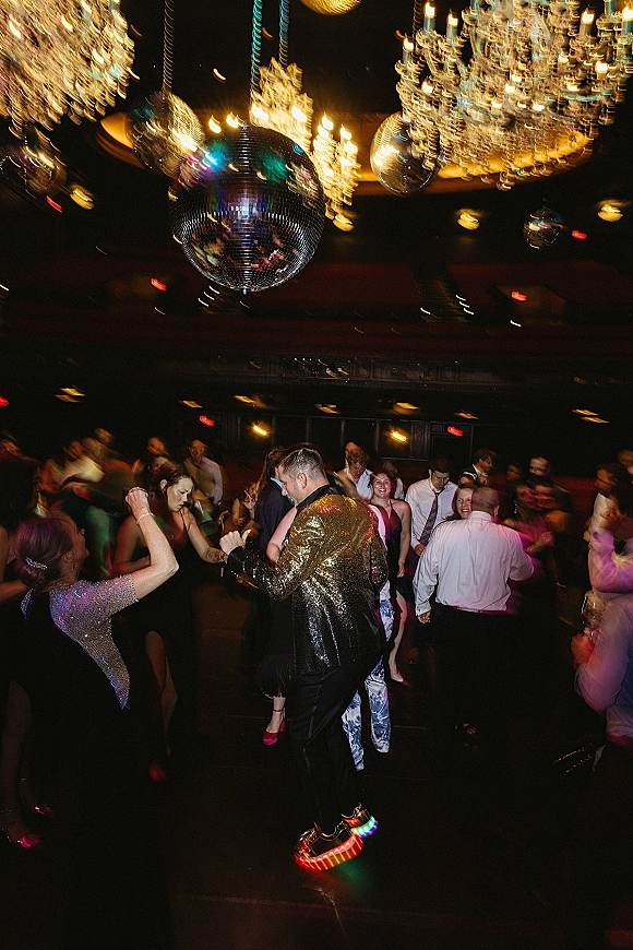 Wedding dance floor packed with guests dancing under chandeliers and disco balls, a groom in a gold sequin jacket in a dark reception room