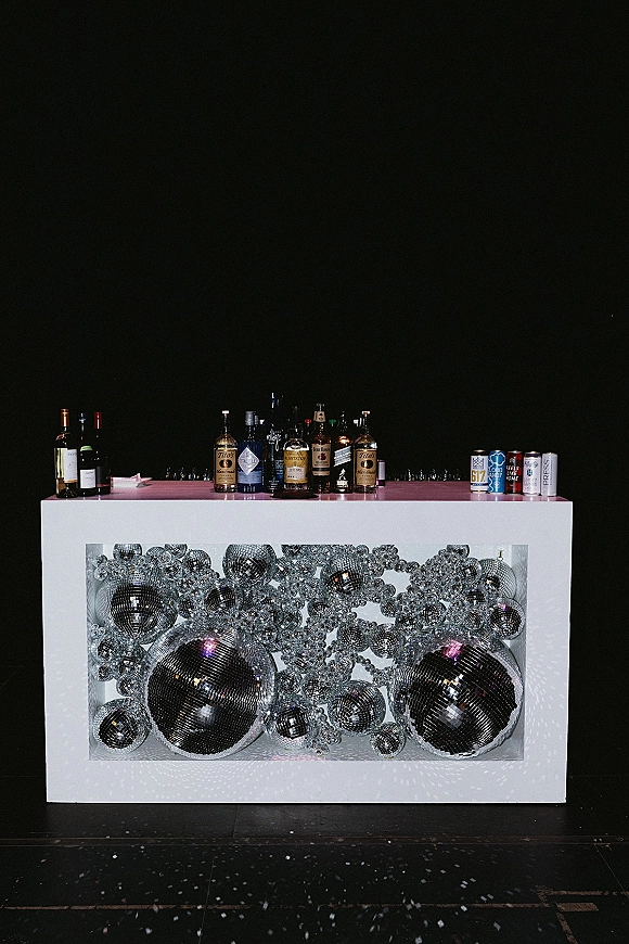 Wedding bar setup with a bar counter lined with liquor, wine and canned drinks, plus disco balls and mirrored ornaments against a black wall floor with confetti