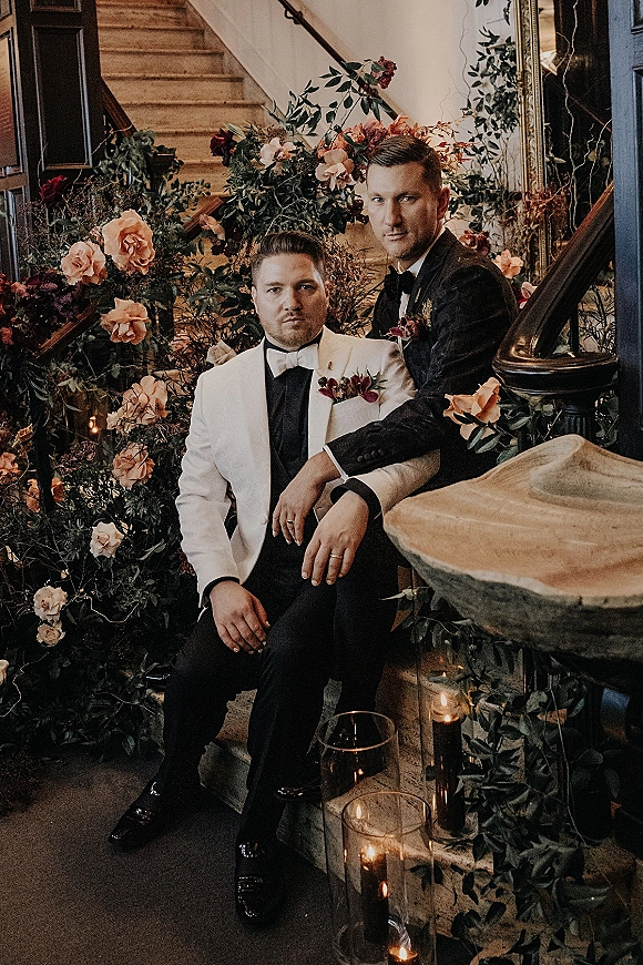 Couple portrait of two grooms in black tux and white dinner jacket with bow ties, on an indoor staircase amid roses and candles