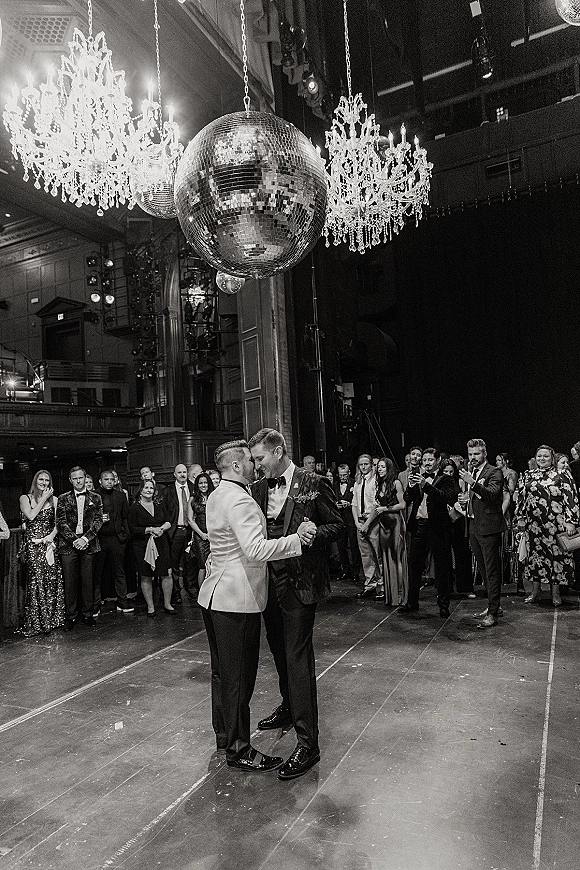 First dance of two grooms in tuxedos during a same sex wedding first dance beneath a disco ball on a theater stage with guests watching