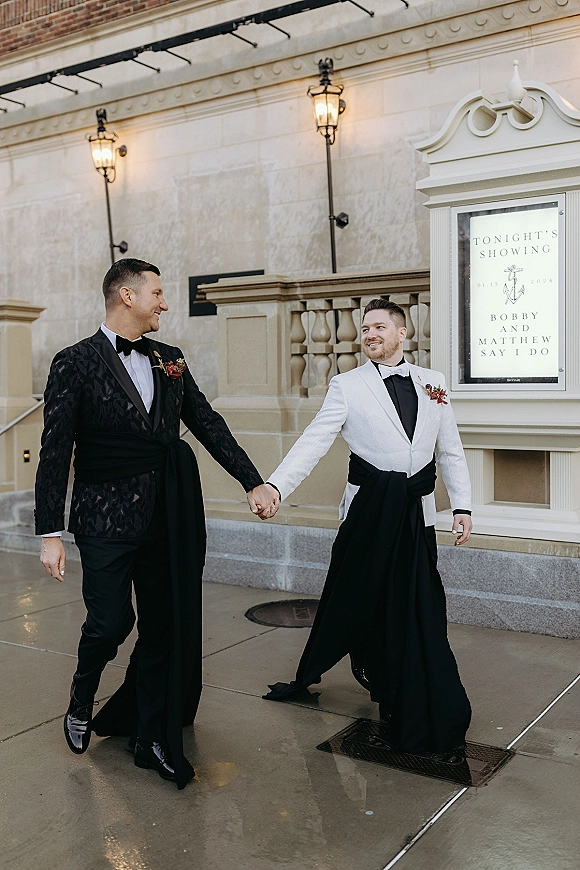 Couple portrait of grooms holding hands in tuxedos with boutonnières, walking beneath a theater marquee on a city sidewalk