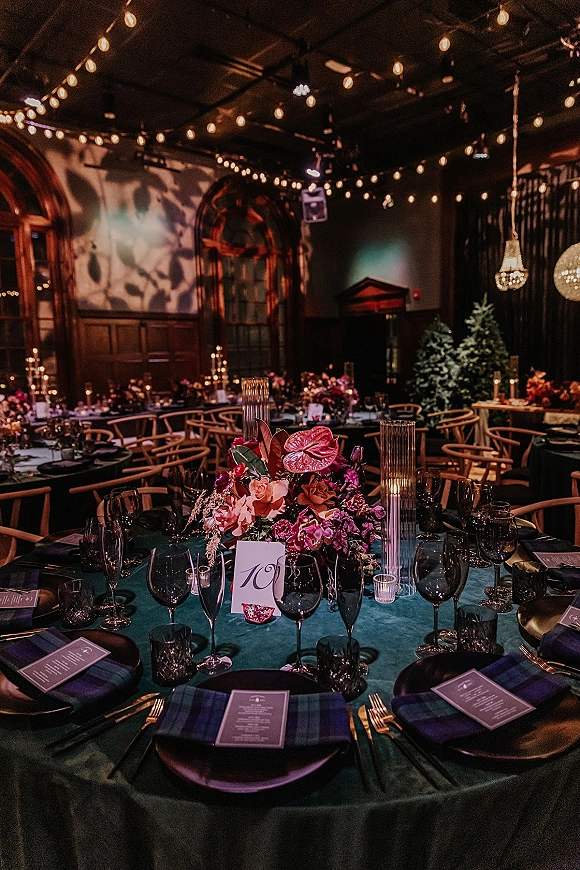 Reception tablescape with a wedding table centerpiece of anthurium and roses, taper candles, and place cards under string lights in a dark hall