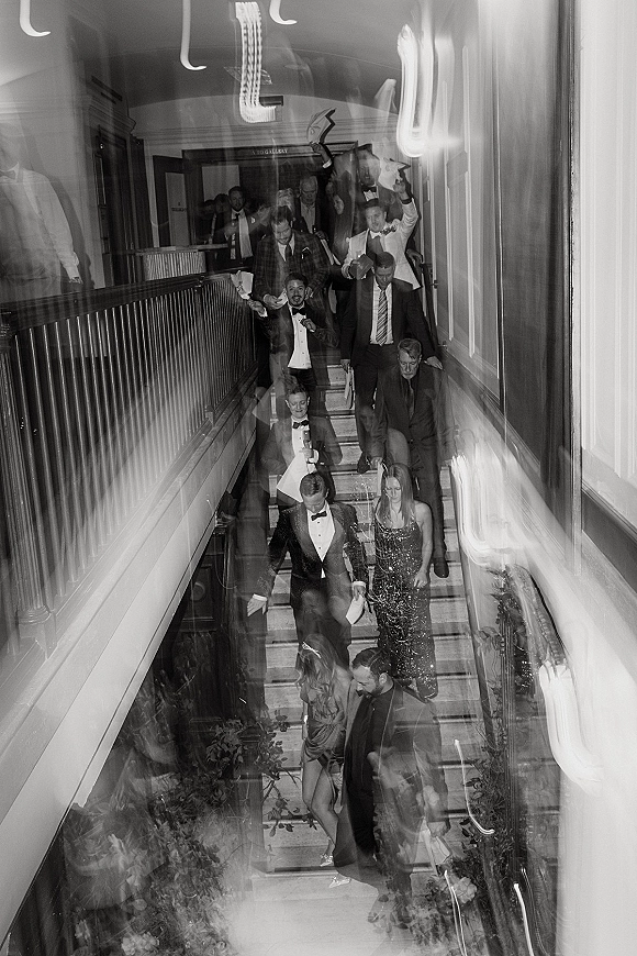 Wedding exit with tuxedoed guests and formal gowns cheering on a venue staircase, one holding a drink under warm wall lights