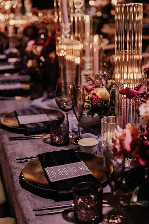 Reception tablescape with candlelit wedding table styling, gold charger plates, black napkins, floral centerpiece, and dim bokeh lighting