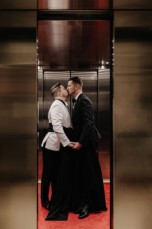 Wedding kiss portrait of two grooms in black tie, one in a white dinner jacket, sharing a kiss by metallic elevator doors on a red carpet